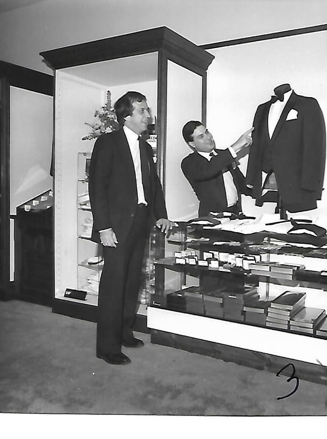 A black-and-white photo of the first shop: two men in suits beside a glass case, one pointing to a tuxedo on a display form