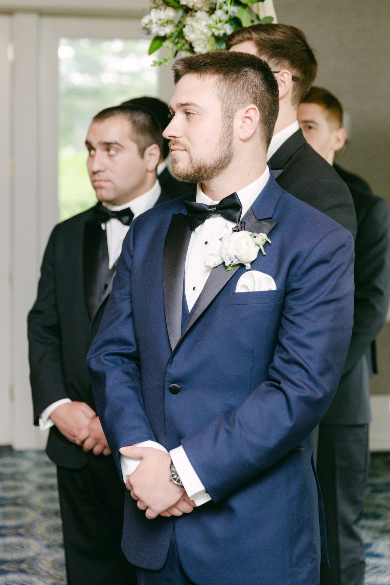 A groom in a blue tuxedo with a white boutonniere standing with his groomsmen