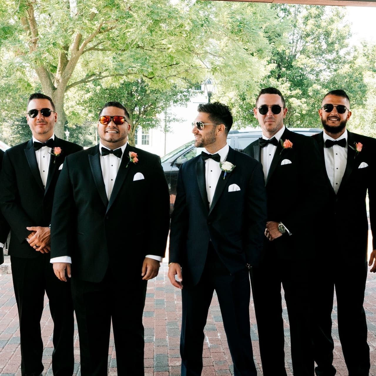 Five groomsmen in black tuxedos and sunglasses standing together outdoors