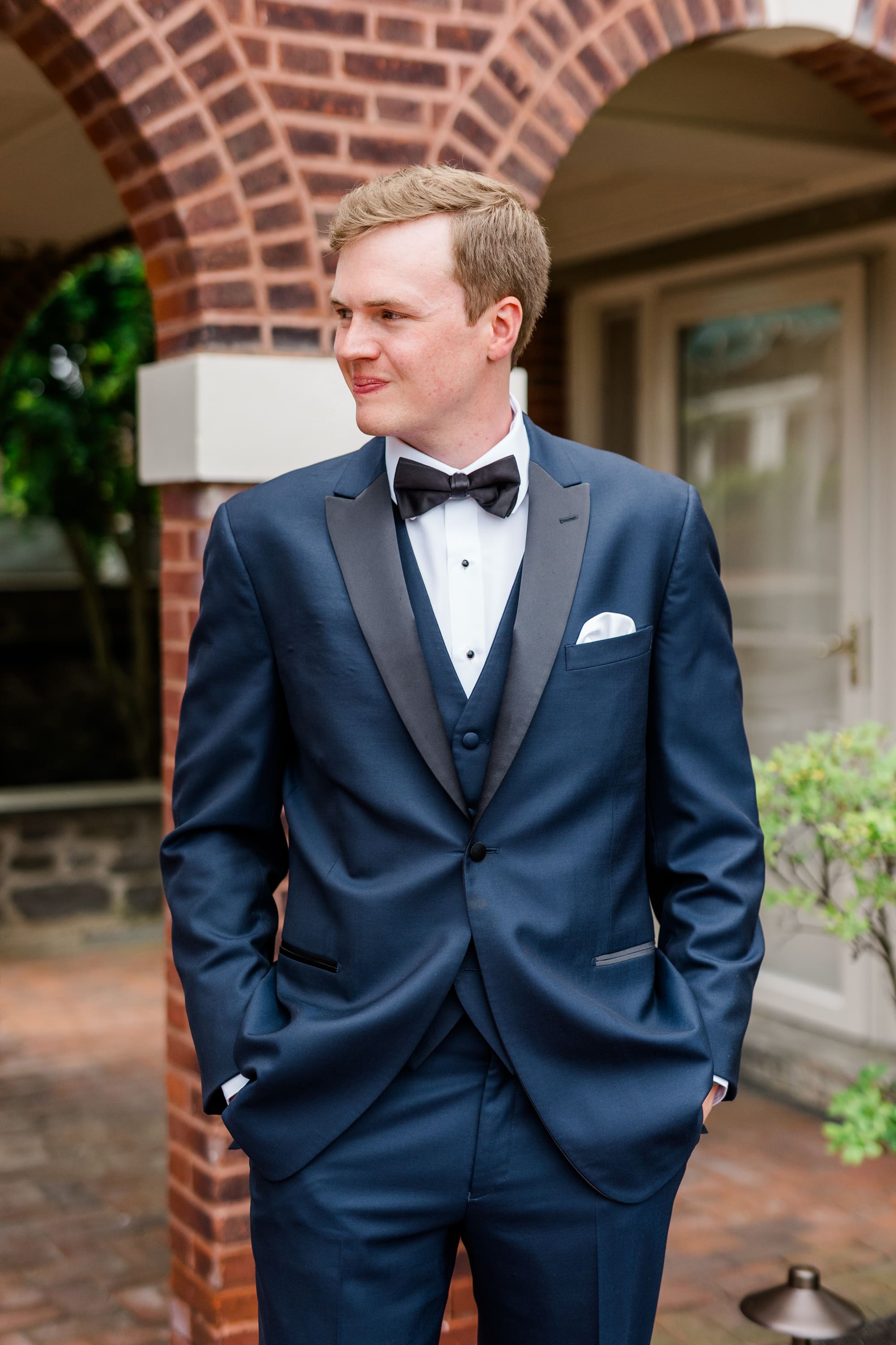 A groom in a navy shawl-collar tuxedo and bow tie standing under a brick archway