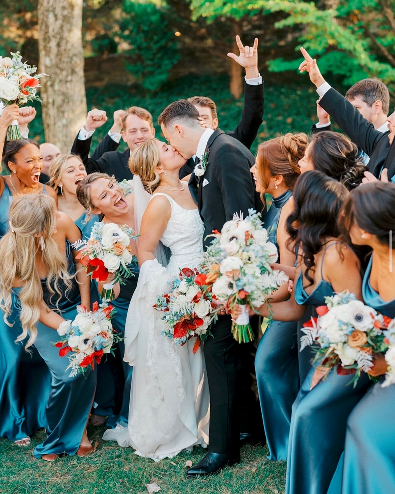 A wedding party celebrating on a lawn, the groom in a black tuxedo kissing the bride among bridesmaids in teal