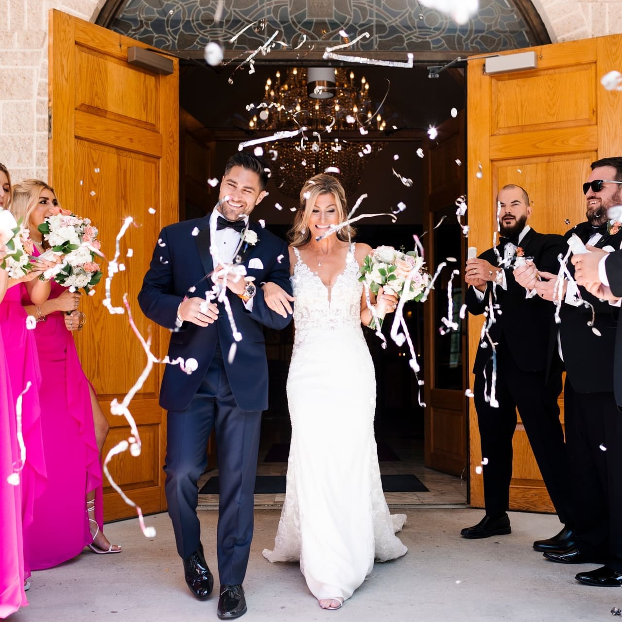 A bride and groom in a navy tuxedo leaving a church through a shower of streamers, groomsmen cheering