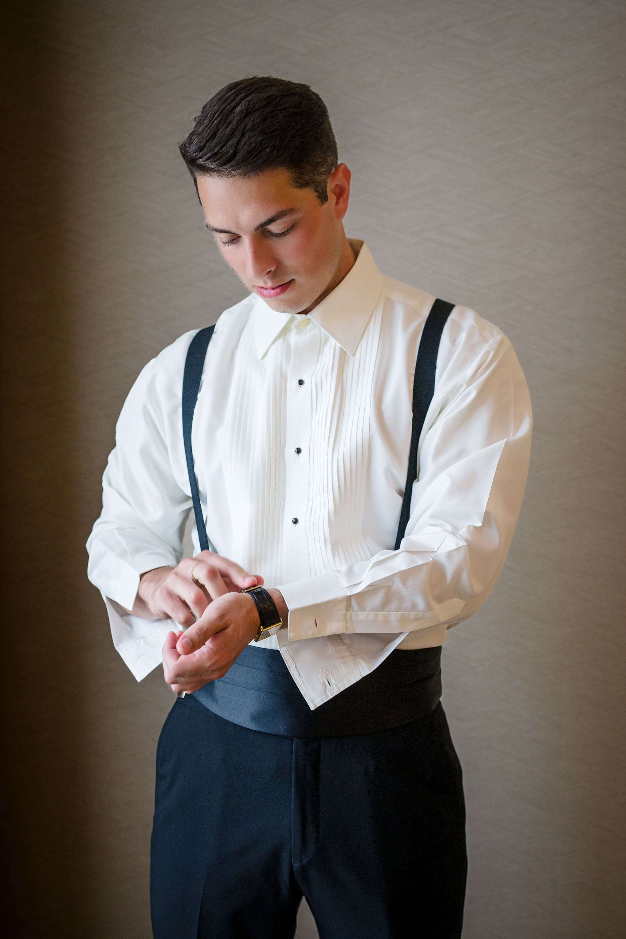 A groom in a pleated tuxedo shirt, suspenders and cummerbund fastening his cuff