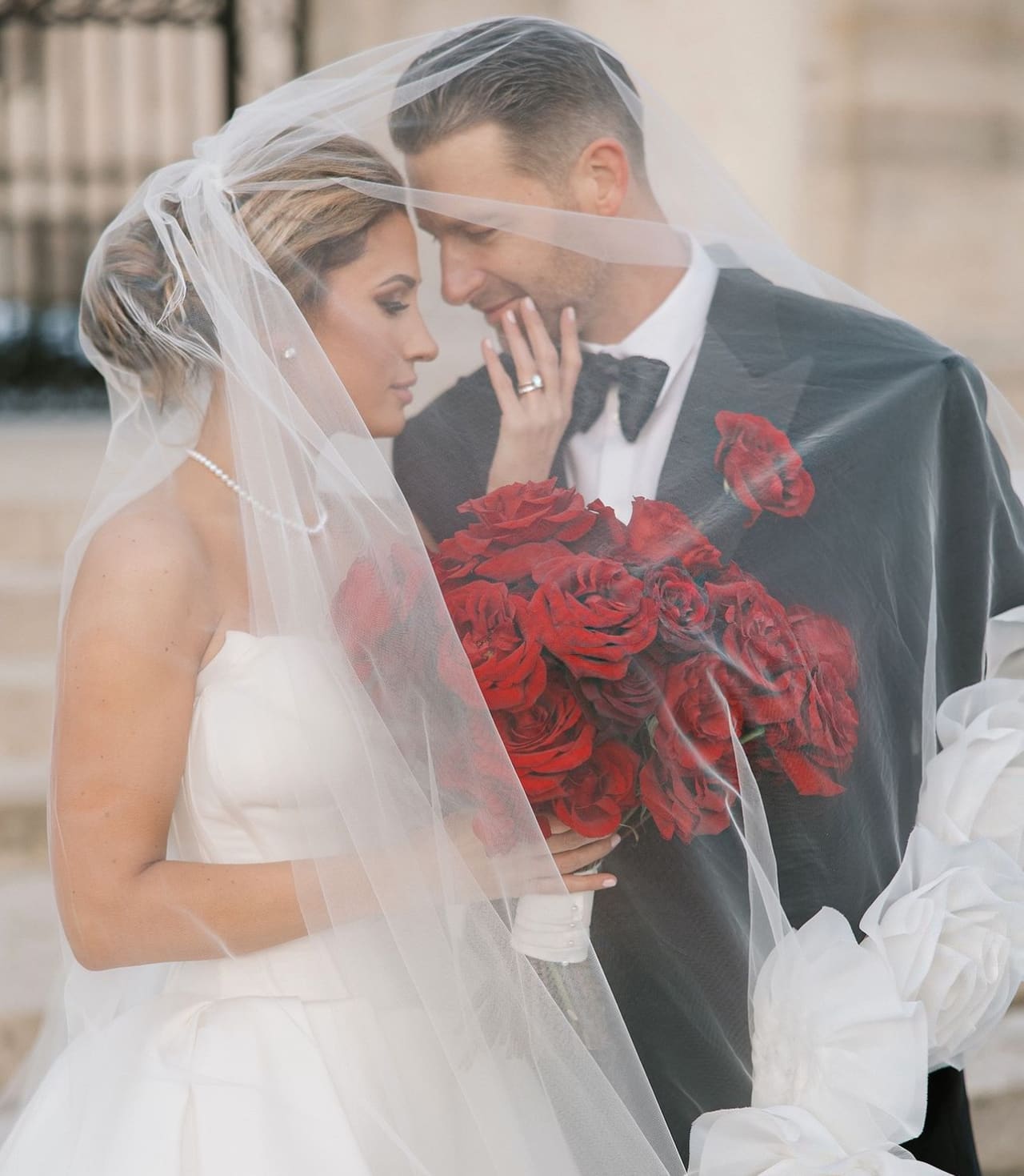 A groom in a black tuxedo and a bride holding red roses, together under her veil