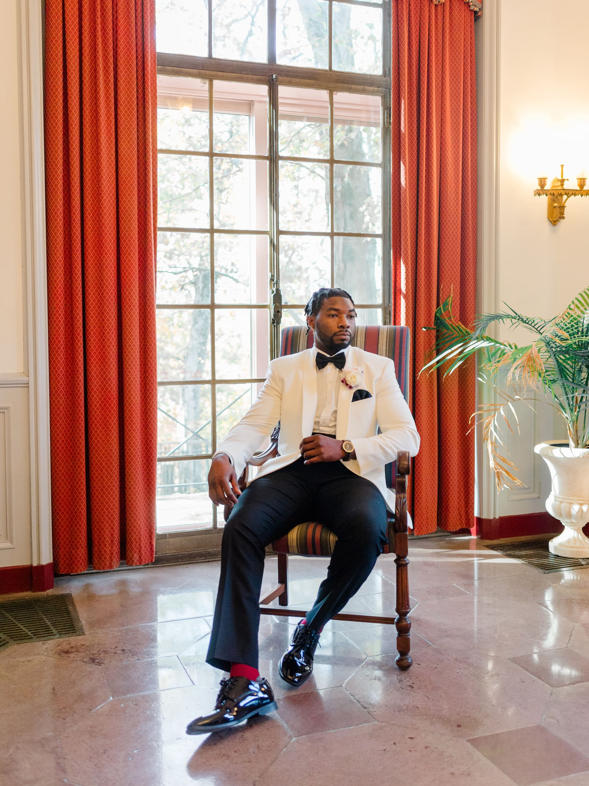 A man in a white dinner jacket and black trousers seated in a sunlit hall with tall red curtains