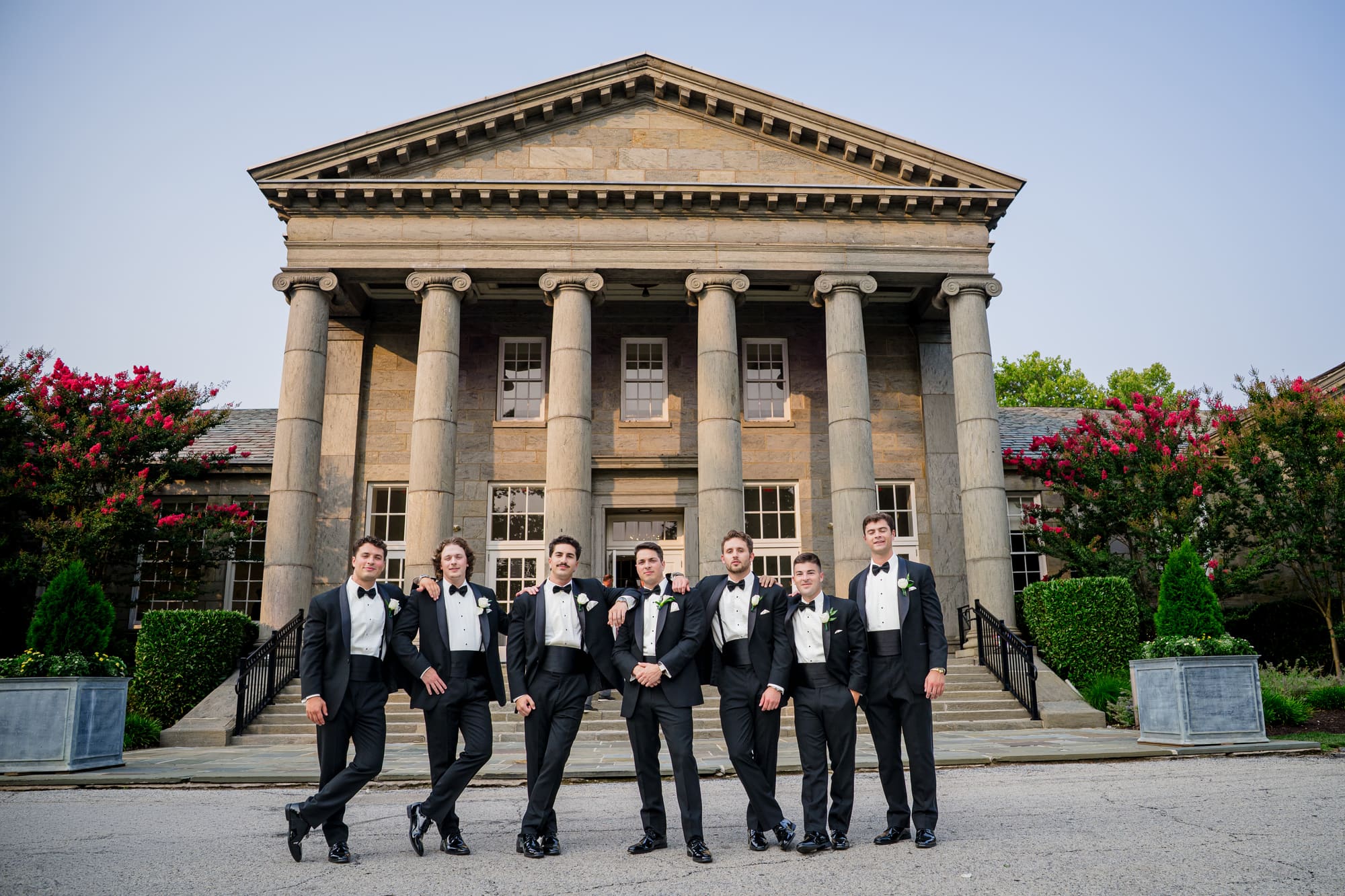 Seven groomsmen in black tuxedos posing in front of a columned stone building