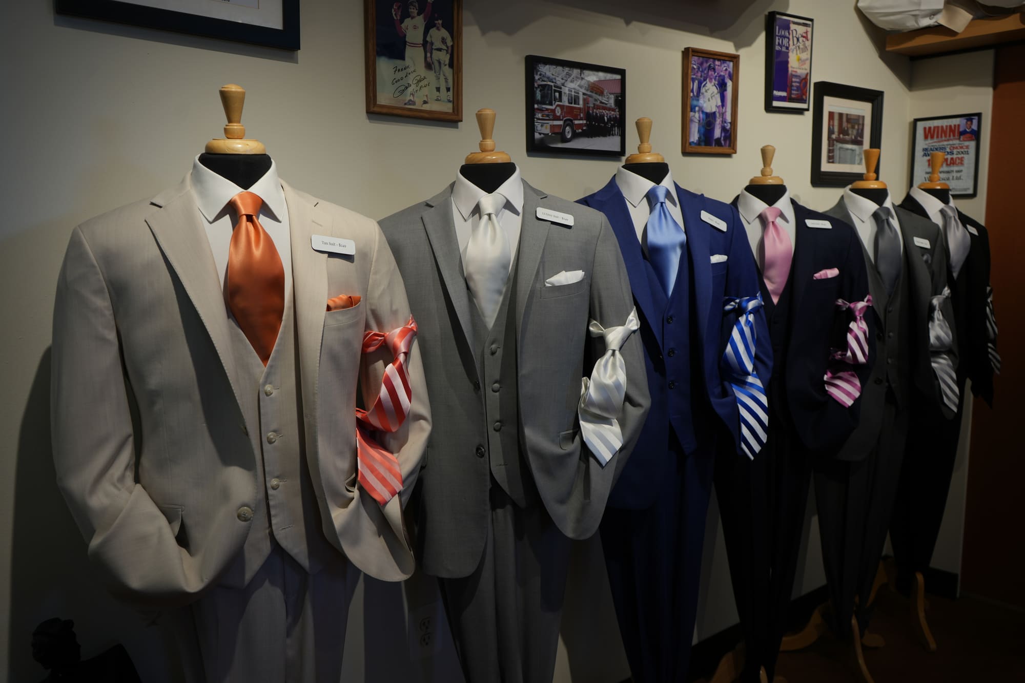 The rental lineup on display forms: light grey and charcoal suits, a navy tuxedo and black tuxedos, with ties in orange, silver, blue and pink