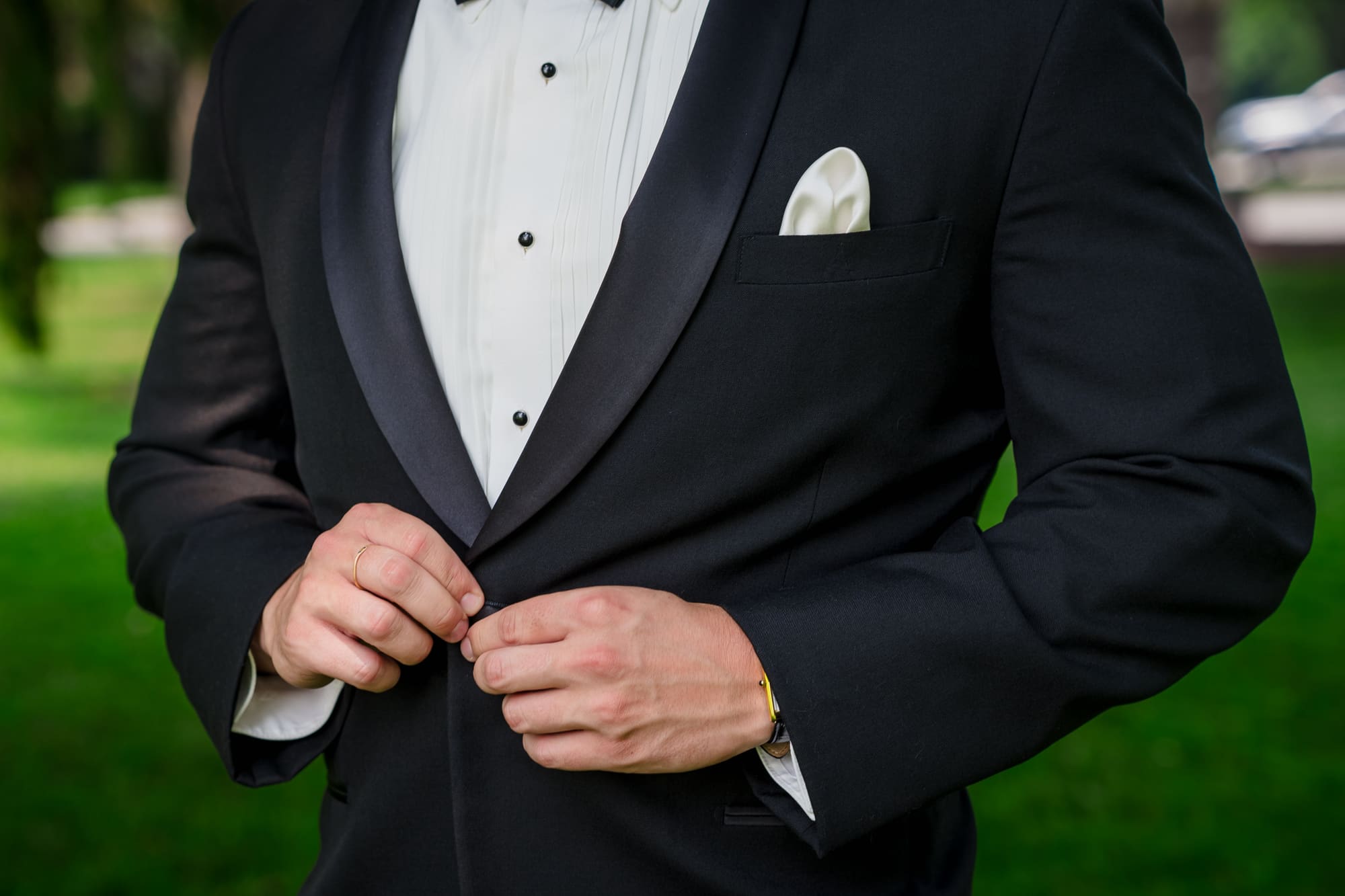 Close-up of a groom buttoning a black shawl-collar tuxedo jacket over a pleated shirt with studs