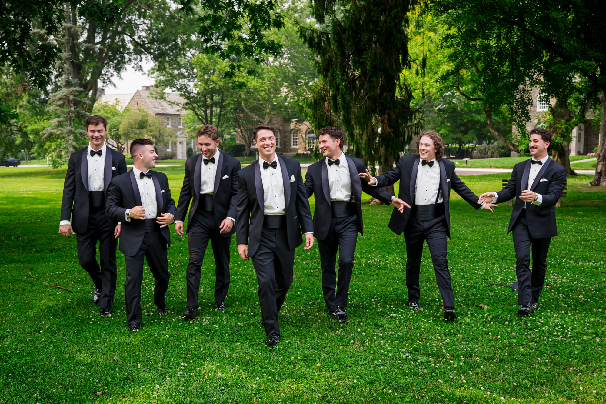 A groom and six groomsmen in black tuxedos walking across a lawn together, laughing